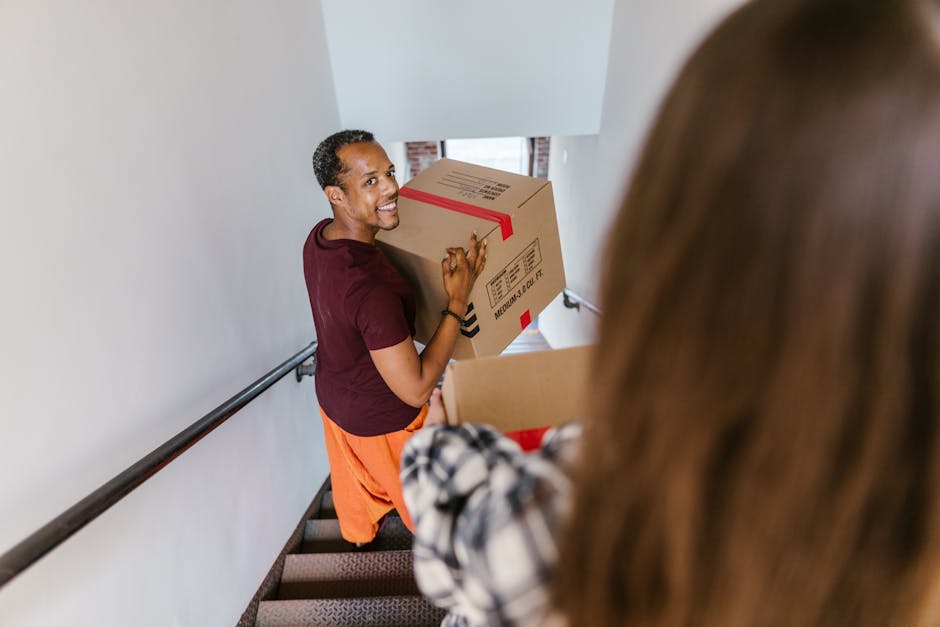 A man with dark skin, short curly hair, and wearing a maroon t-shirt and orange trousers is smiling while holding a medium-sized cardboard box sealed with red packing tape. He is standing on a stairca